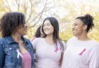 Three women wearing pink shirts with pink cancer bows attached smiling at each other.