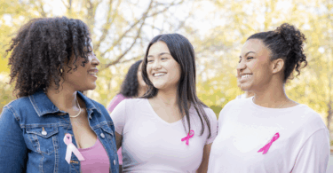 Three women wearing pink shirts with pink cancer bows attached smiling at each other.