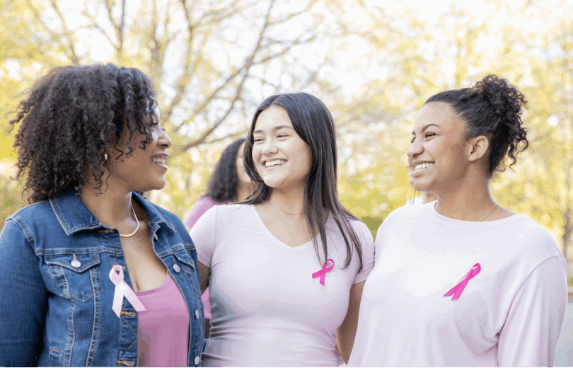Three women wearing pink shirts with pink cancer bows attached smiling at each other.