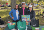 A man and two women stand together in the park in front of a table with goodie bags.