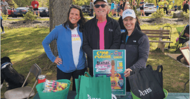A man and two women stand together in the park in front of a table with goodie bags.