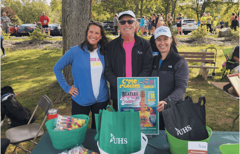 A man and two women stand together in the park in front of a table with goodie bags.